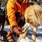 Person in orange jacket and brown gloves giving dog Rigby beef pouch in the snow