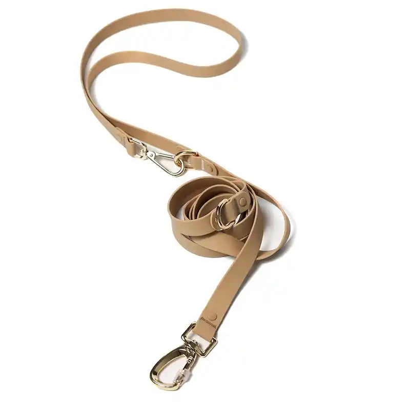 A sand colored silicone coated webbing dog leash with a gold metal D-ring and a buckle, designed for hands-free crossbody wear.