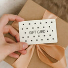 Hand holding a MiAmore gift card over a wrapped present with a beige ribbon.